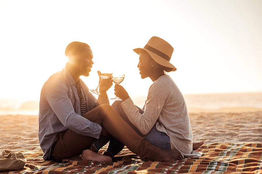 Personal Insurance - Portrait of a Cheerful Young Couple Sitting on a Blanket on the Beach During Sunset While Enjoying a Glass of Wine Together